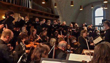 Orchester und Chor treten in einer Kirche auf, maskiertes Publikum sichtbar.